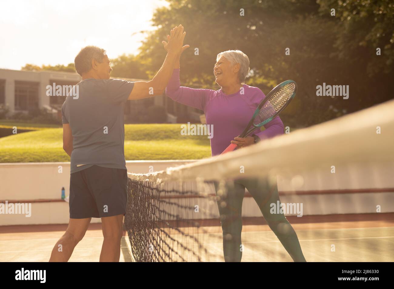 Fröhliches Senioren-Paar mit Biracial, das bei Sonnenuntergang auf dem Tennisplatz Tennis spielte und fünf Punkte gab Stockfoto