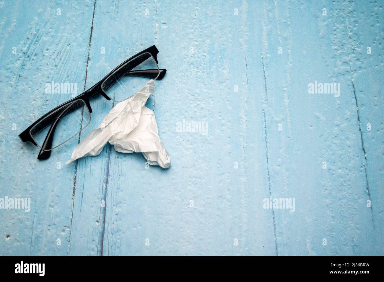 Draufsicht auf Lesebrille mit schwarzen Bügeln und zerknittertem Brillenreinigungstuch auf hellblau lackierter Holzoberfläche Stockfoto