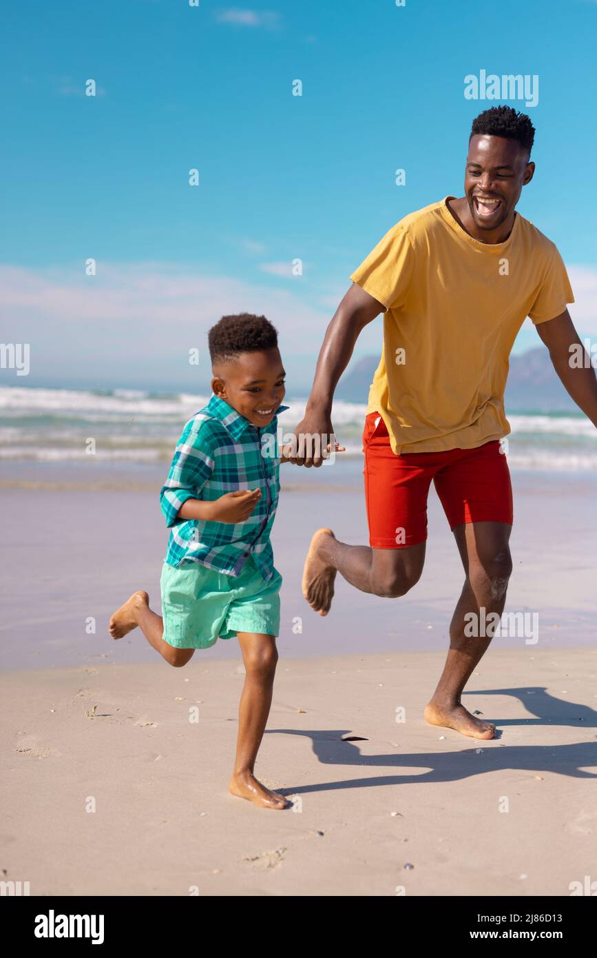 Verspielter afroamerikanischer Junge, der die Hände des jungen Vaters hält und am Strand gegen den blauen Himmel läuft Stockfoto