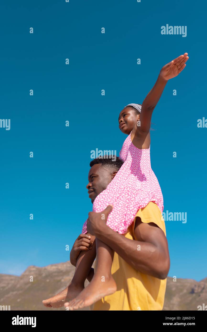 Low-Angle-Ansicht des afroamerikanischen Mädchens mit ausgestreckten Armen, die auf den Schultern des jungen Vaters sitzen Stockfoto