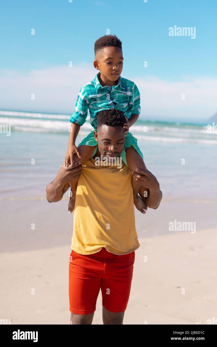afroamerikanischer junger Vater, der seinen Sohn auf den Schultern trägt, während er gegen das Meer und den blauen Himmel steht Stockfoto