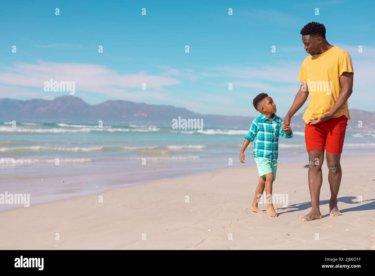 afroamerikanischer Junge, der die Hände des jungen Vaters hält, während er am Strand gegen den blauen Himmel auf Sand läuft Stockfoto