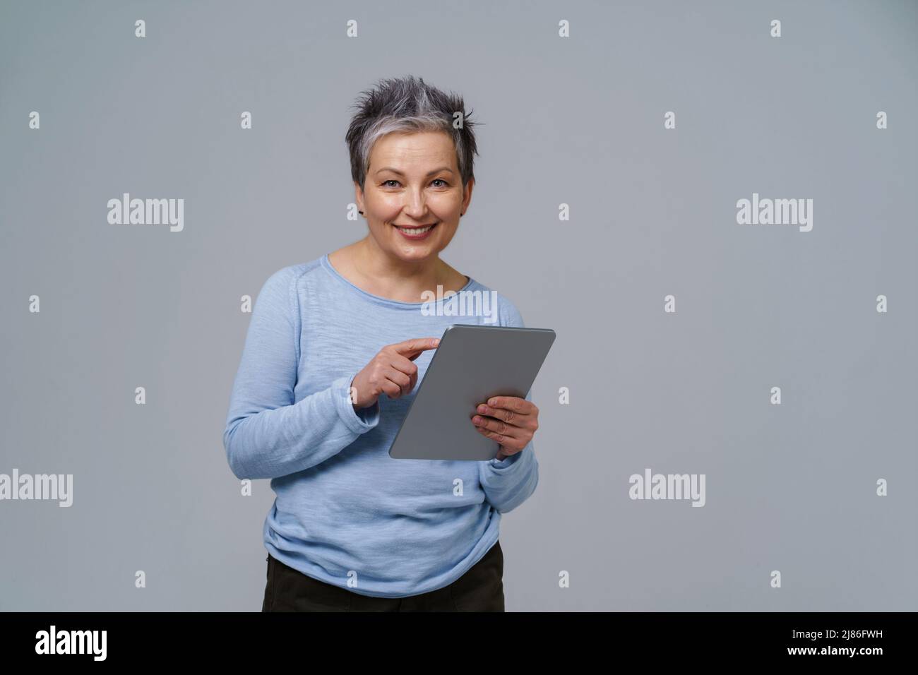 Lächelnde reife grauhaarige Frau 50s hält digitales Tablet bei der Arbeit oder beim Prüfen in sozialen Medien. Hübsche Frau im Jahr 50s in blauer Bluse isoliert auf weiß. Ältere Menschen und Technologien. Getöntes Bild. Stockfoto