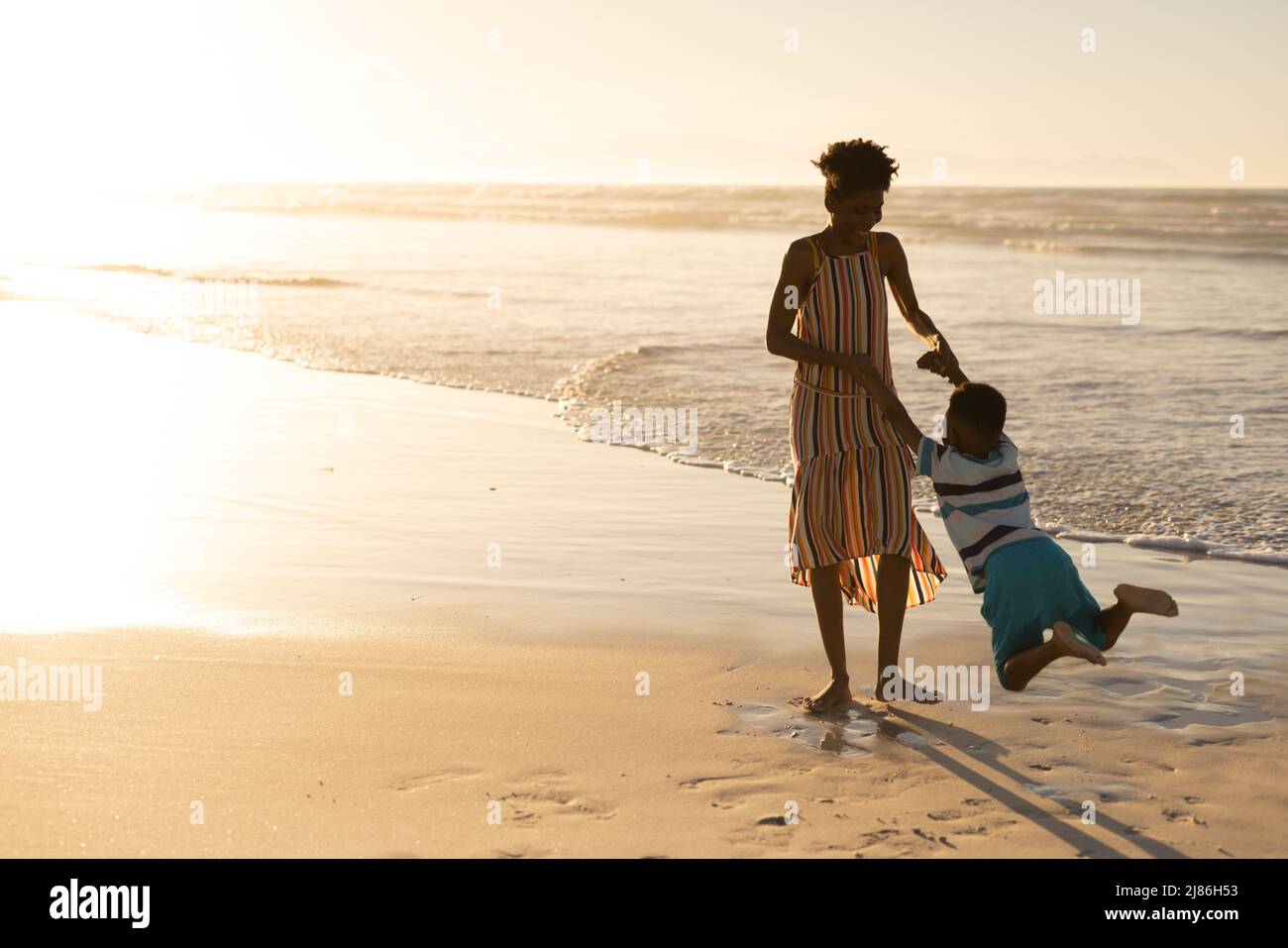 Verspielte afroamerikanische junge Frau, die die Hände des Sohnes hält und ihn am Strand gegen den Himmel dreht Stockfoto