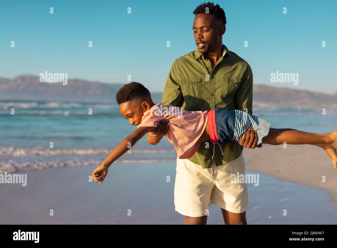 afroamerikanischer junger Vater trägt verspielten Sohn am Strand gegen klaren Himmel während des Sonnenuntergangs Stockfoto