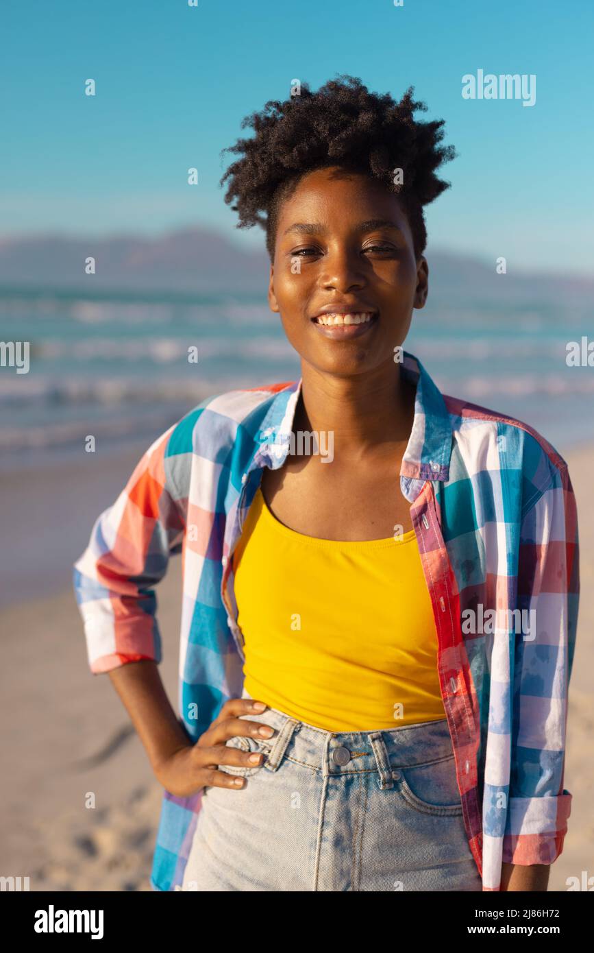 Porträt einer glücklichen afroamerikanischen jungen Frau mit kurzen Haaren und Hand auf der Hüfte am Strand gegen den Himmel Stockfoto