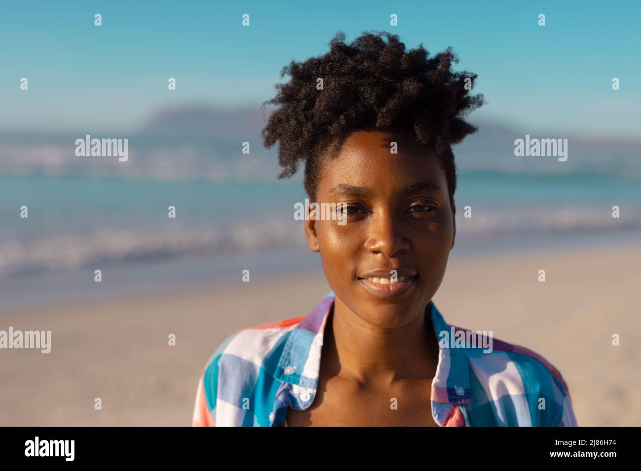 Porträt einer lächelnden afroamerikanischen jungen Frau mit kurzen lockigen Haaren am Strand vor klarem Himmel Stockfoto