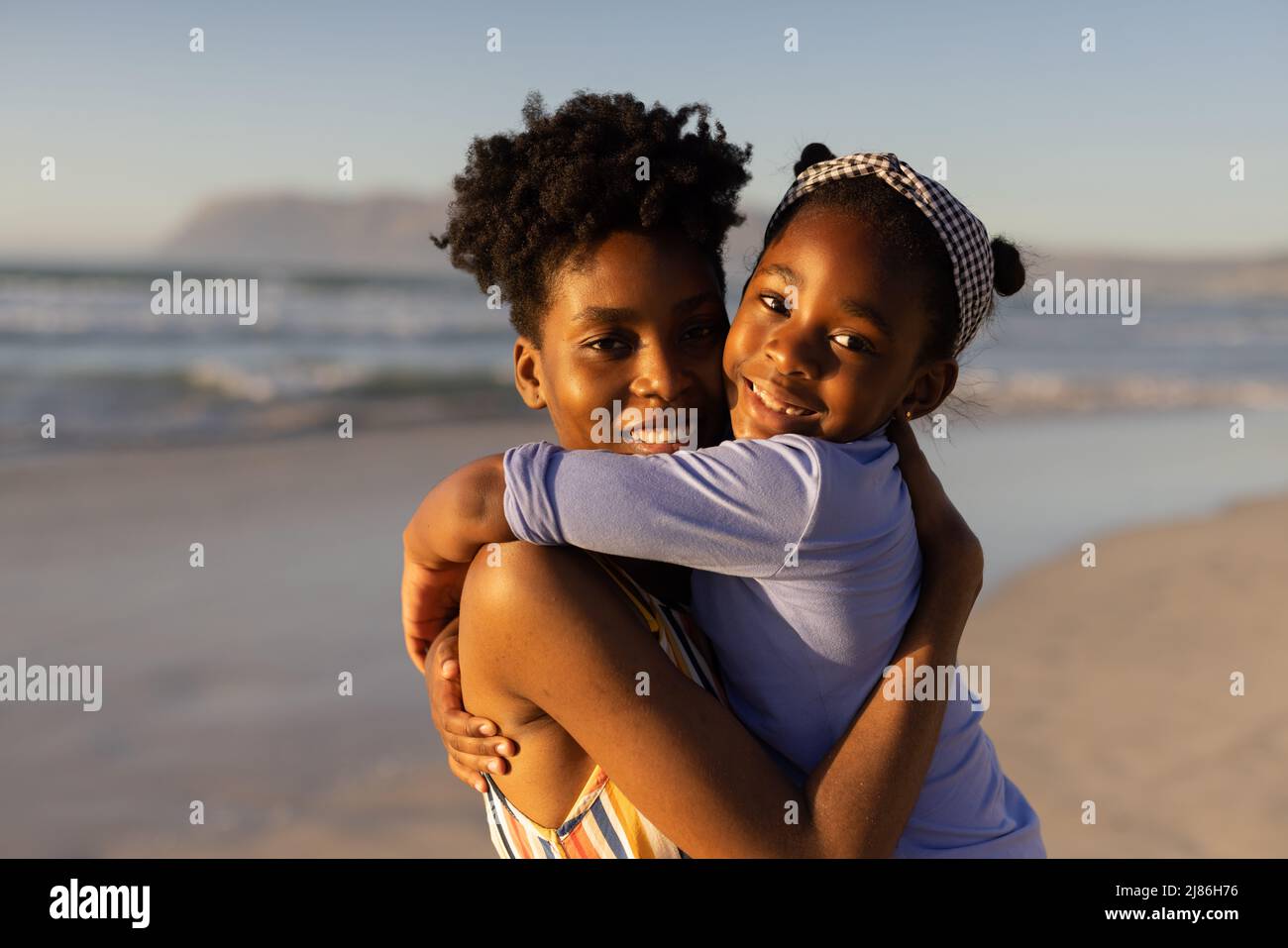 Porträt einer lächelnden afroamerikanischen jungen Frau mit kurzen Haaren, die ihre Tochter gegen das Meer umarmt Stockfoto