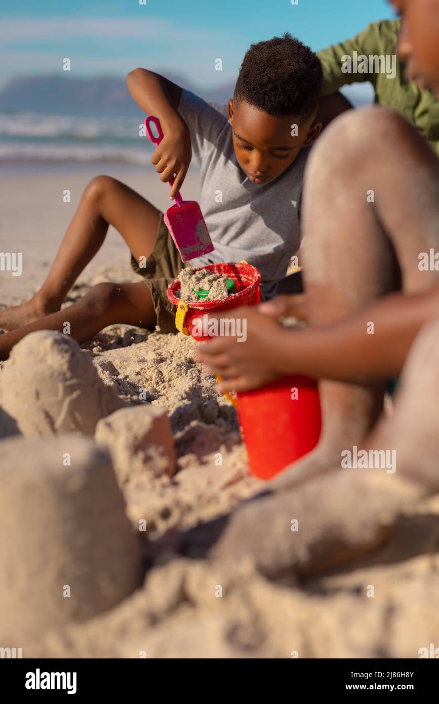 afroamerikanischer Junge, der mit Sand, Eimer und Schaufel spielt, während er mit Vater und Schwester am Strand sitzt Stockfoto