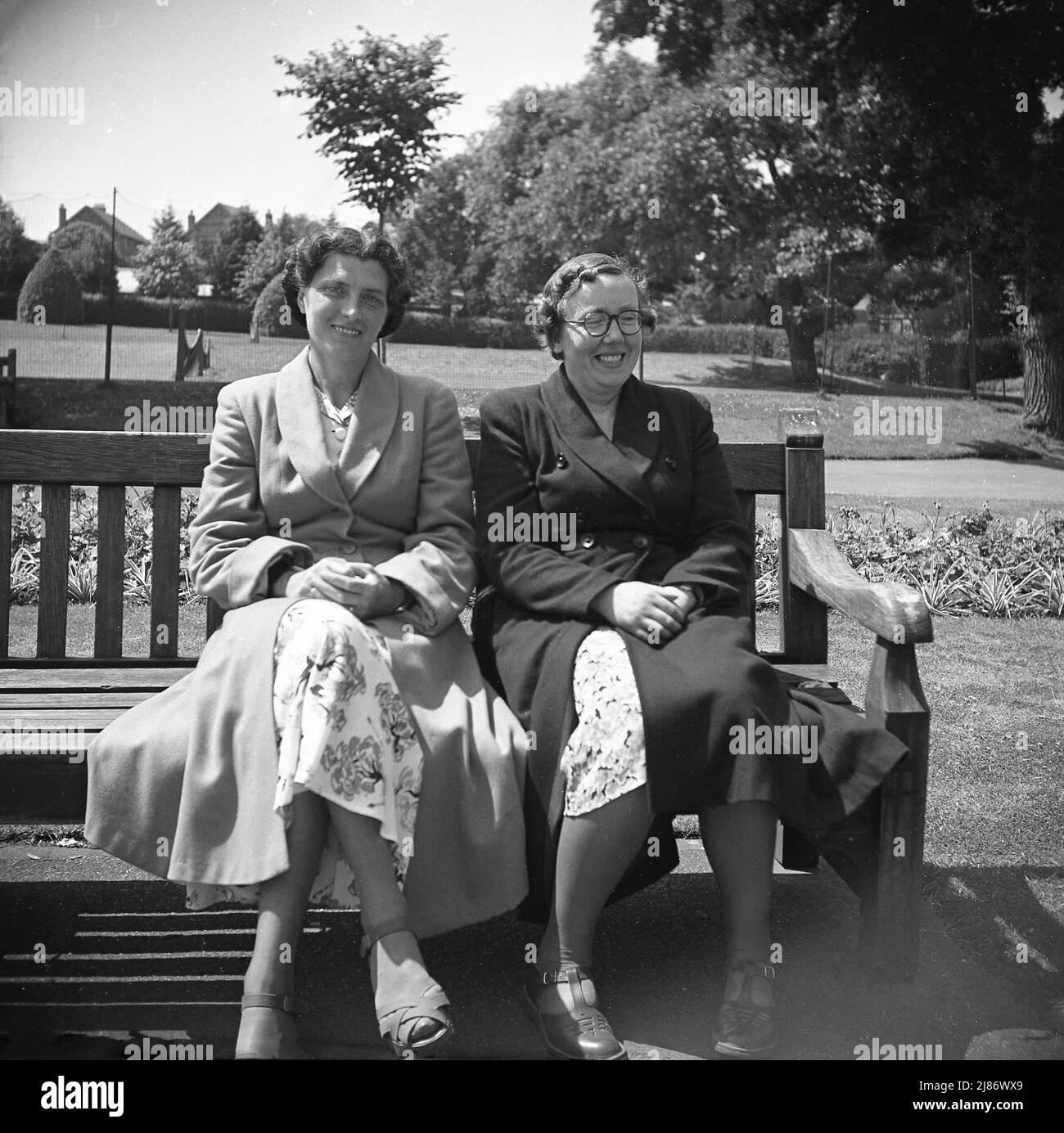 1950s, historisch, zwei Frauen sitzen zusammen auf einer Parkbank an der Küste, mit einem Lachen, Westgate-on-Sea, Margate, Kent, England, Großbritannien. Sie tragen lange Mäntel über ihren Blumenmusterkleidern, gewöhnliche weibliche Kleidung der Ära. Hinter ihnen befinden sich Tennisplätze. Stockfoto