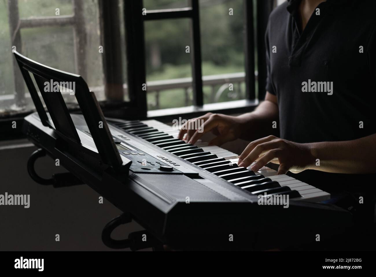 Junger latein, der zu Hause am Fenster E-Piano oder Synthesizer spielt, Moment der Entspannung und Reflexion mit Musik, um sich zu unterhalten Stockfoto