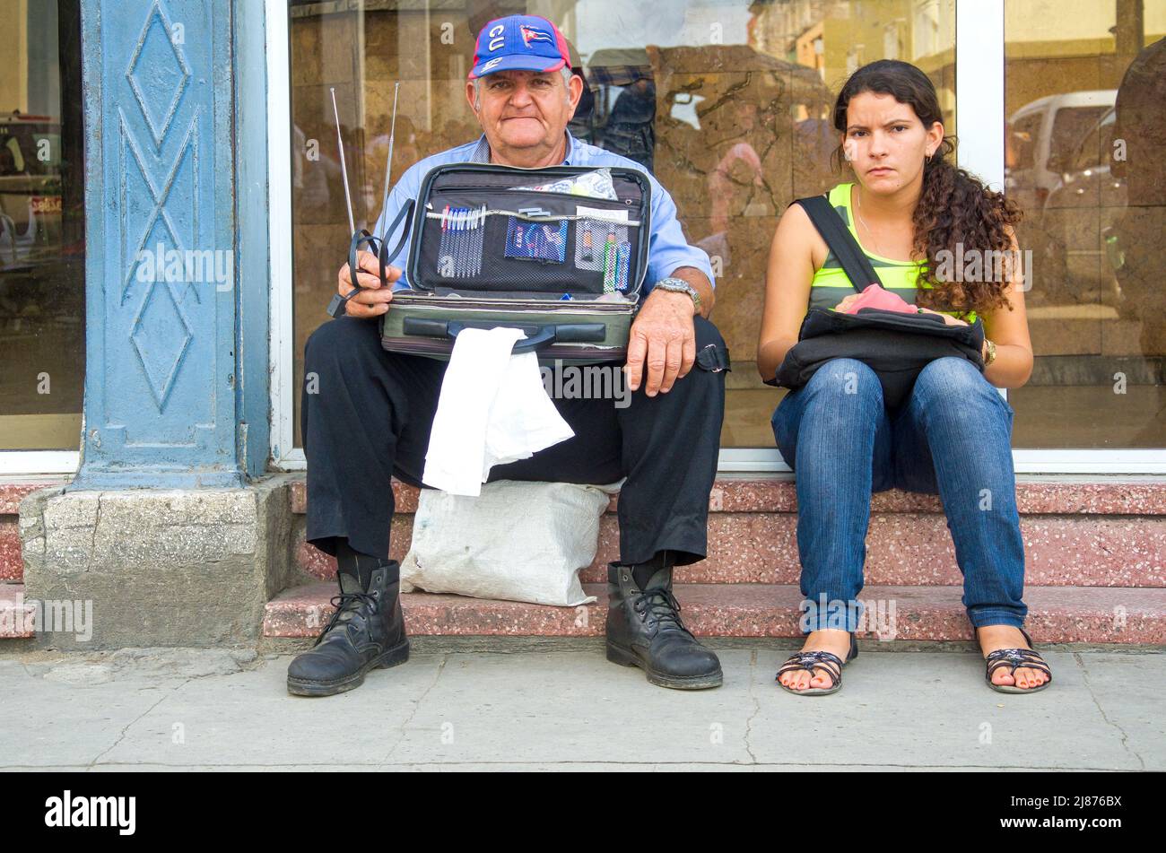 Ein älterer Mann verkauft kleine Gegenstände, die in der Post in der Innenstadt sitzen. Er trägt eine Baseballmütze mit kubanischen Symbolen. Eine junge Frau sitzt an seiner Seite. Stockfoto