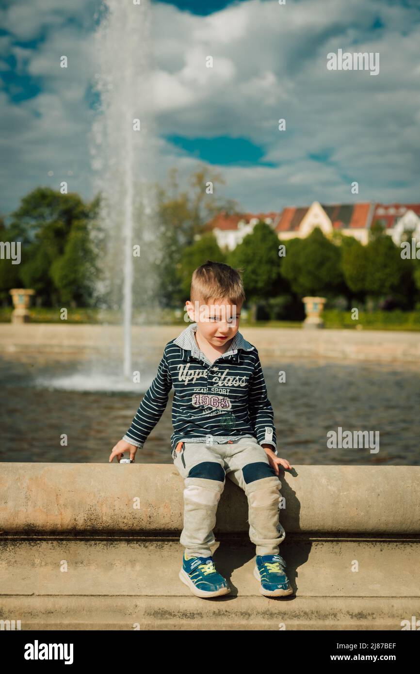 Ein kleiner Junge sitzt an einem sonnigen Tag in der Nähe eines Brunnens. Stockfoto
