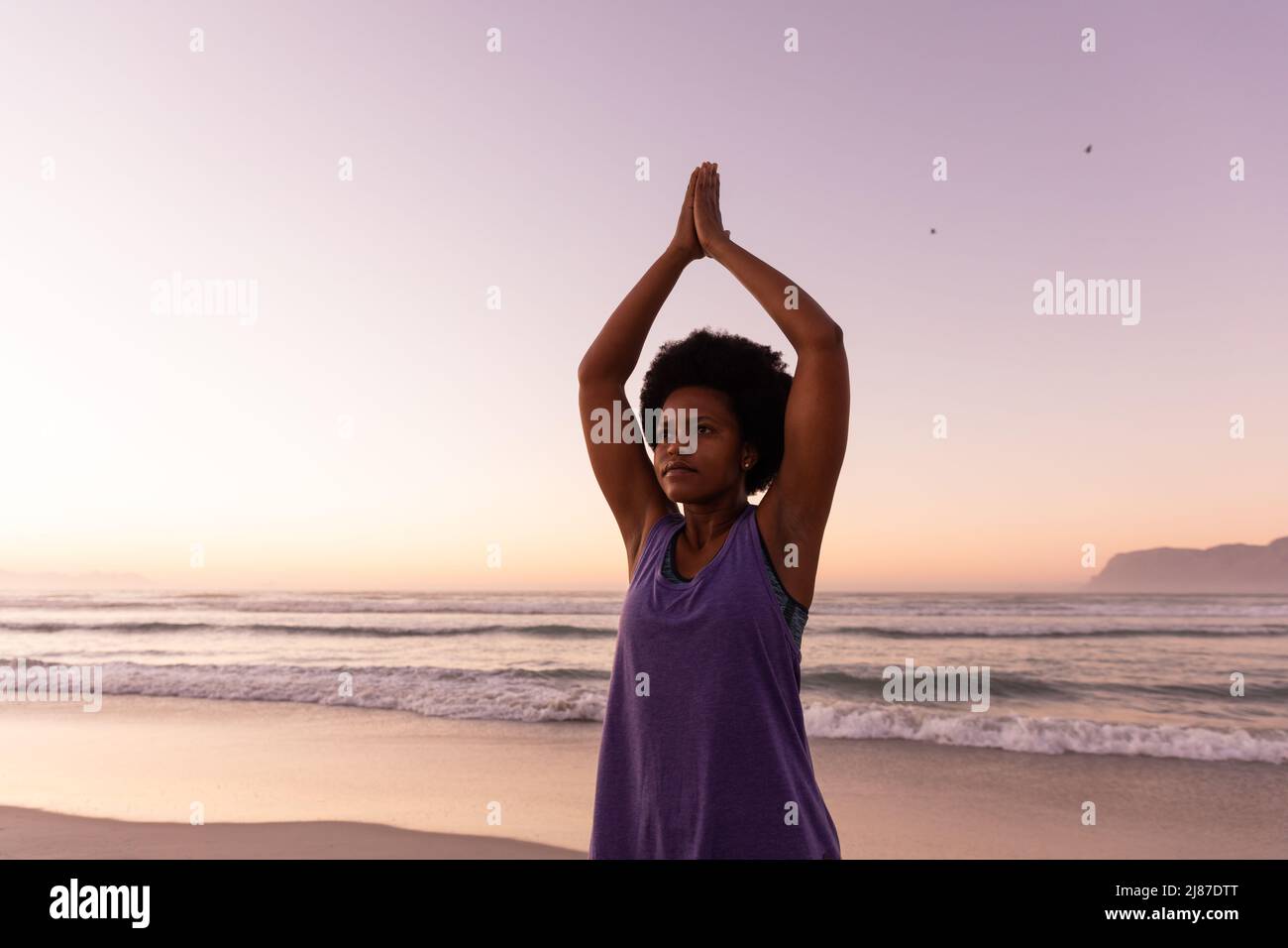 afroamerikanische reife Frau mit erhobenen Armen, die während des Sonnenuntergangs am Strand gegen den klaren Himmel trainiert Stockfoto