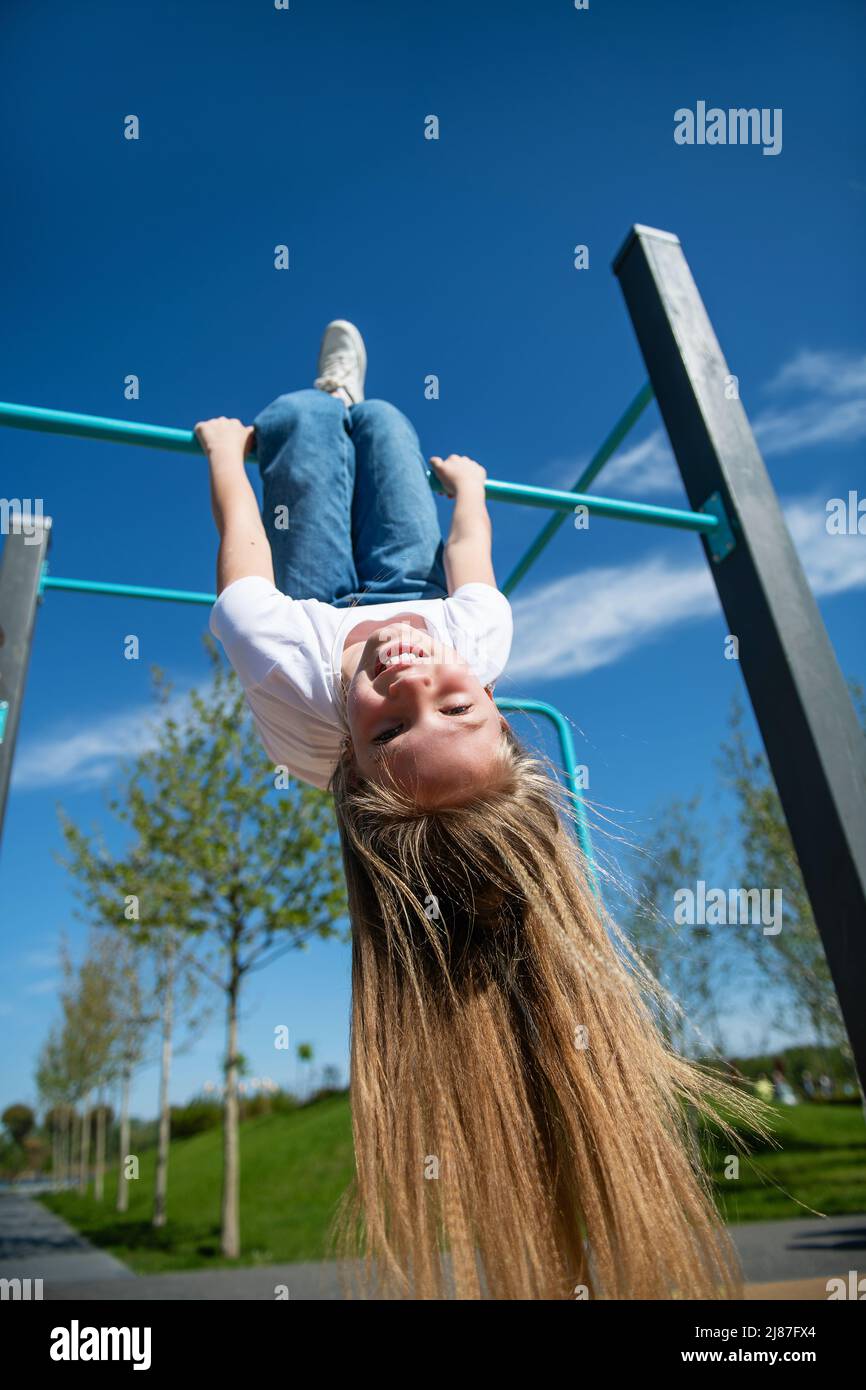 Kleines lustiges Mädchen mit langen Haaren, die kopfüber auf der horizontalen Stange hängen. Ansicht von unten, Weitwinkel. Stockfoto