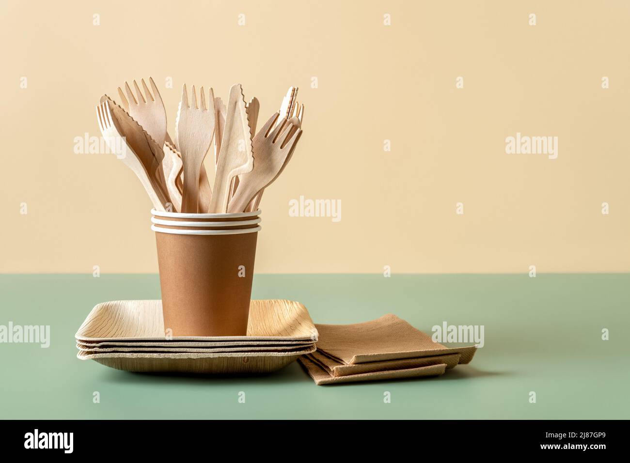 Umweltfreundliches Einweggeschirr auf grün-beigem Hintergrund. Holzgabeln und Messer in Pappbechern auf Bambusplatten. Einmal-Geschirr. Stockfoto