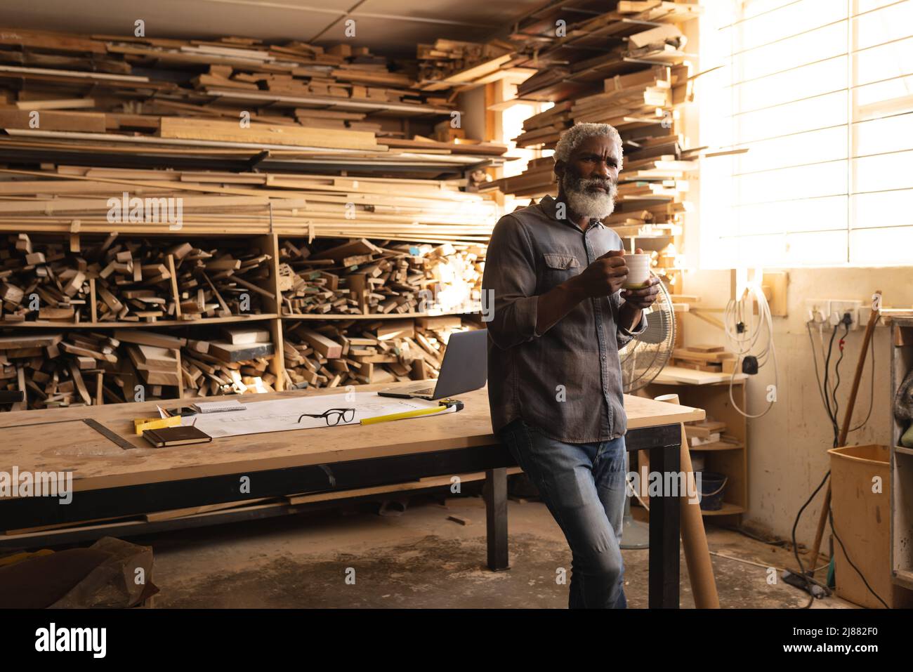 Nachdenklicher afroamerikanischer reifer Zimmermann, der in der Holzwerkstatt Kaffee getrunken hat Stockfoto