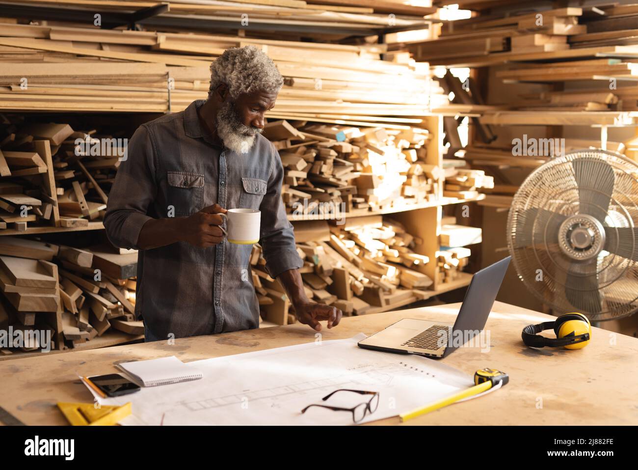 afroamerikanischer reifer Zimmermann, der in der Holzwerkstatt Kaffee am Laptop arbeitete Stockfoto