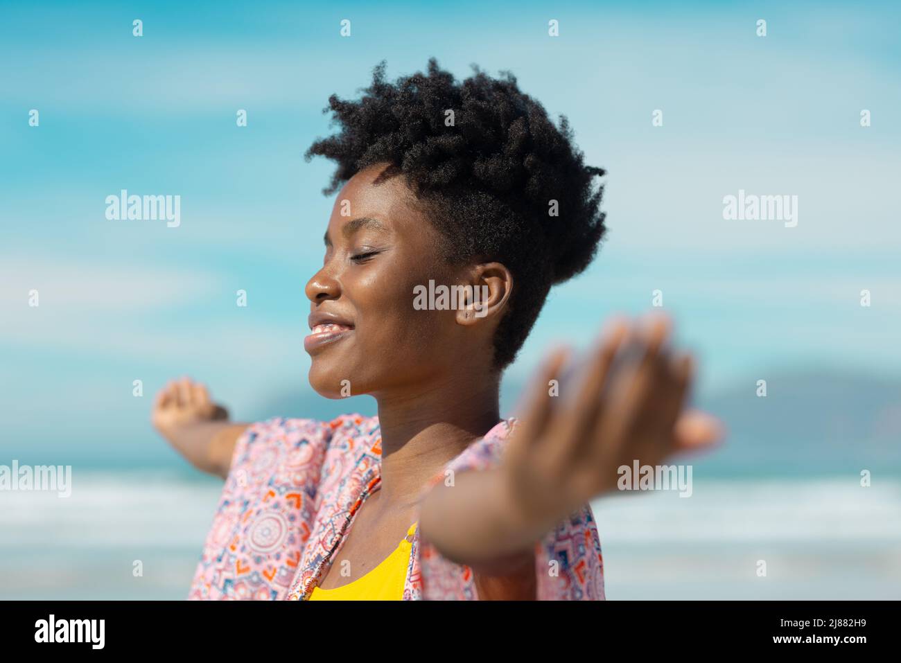 Lächelnde afroamerikanische junge Frau mit geschlossenen Augen und ausgestreckten Armen am sonnigen Tag Stockfoto