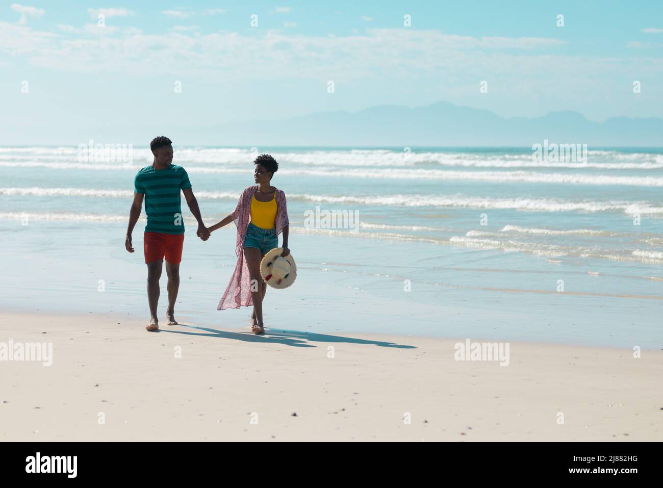 afroamerikanische junge Frau mit kurzen Haaren, die die Hände des Freundes hält, während sie am Strand spazieren geht Stockfoto