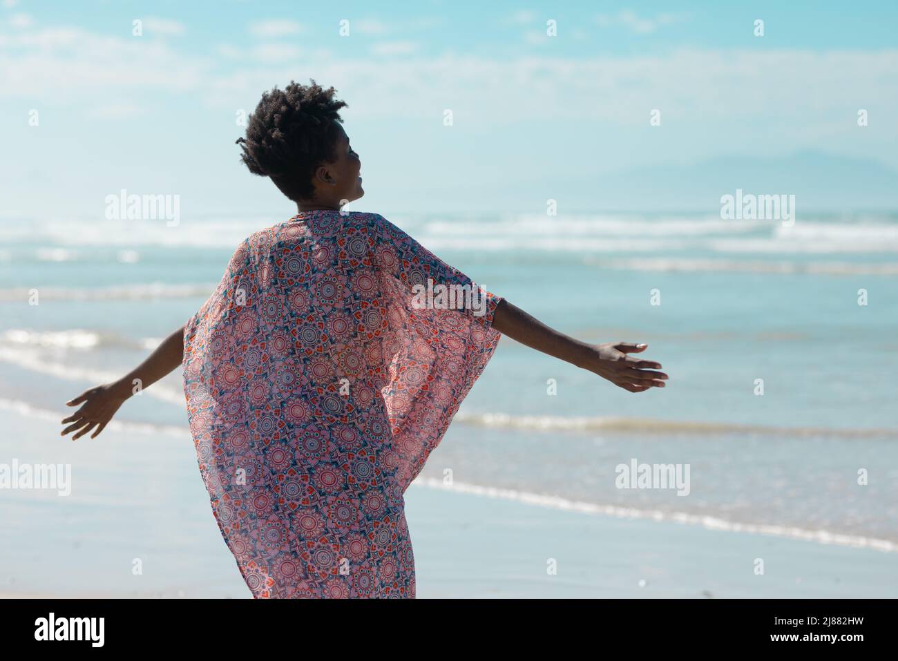 Rückansicht einer afroamerikanischen jungen Frau in Sarong-Jacke mit ausgestreckten Armen am Strand Stockfoto