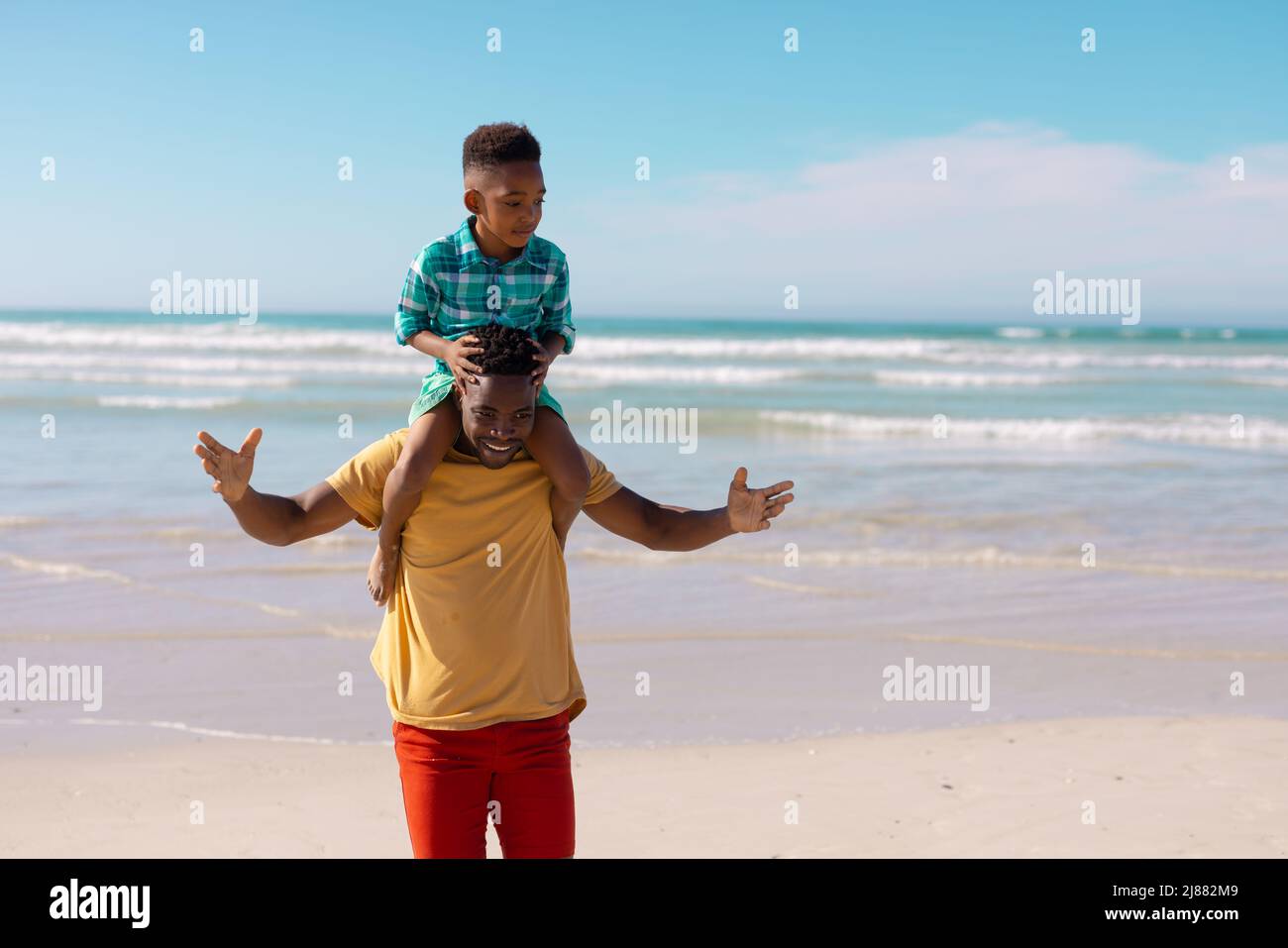 Glücklicher afroamerikanischer junger Mann, der seinen Sohn auf den Schultern trägt, während er gegen Meer und Himmel steht Stockfoto