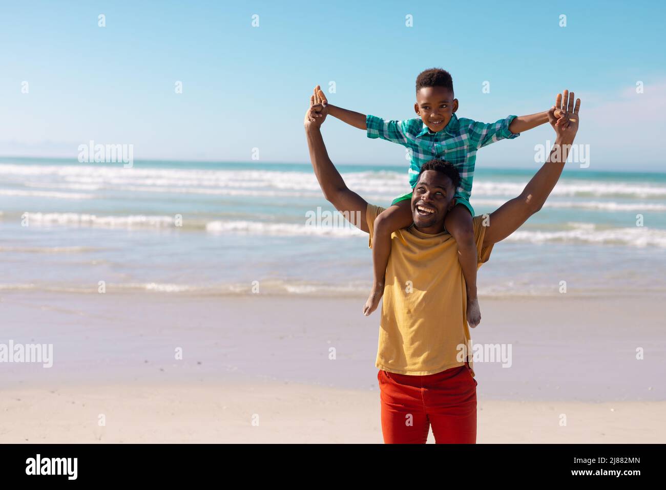 Fröhlicher afroamerikanischer junger Mann, der seinen Sohn auf Schultern trägt und gegen das Meer und den blauen Himmel steht Stockfoto
