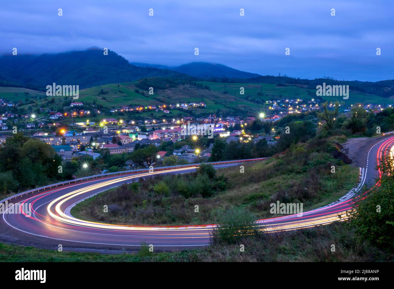 Wolkiger Abend auf einer Bergstraße in den ukrainischen Karpaten. Spuren von Scheinwerfern von sich schnell bewegenden Autos. Kleines Dorf mit Lichtern im val Stockfoto