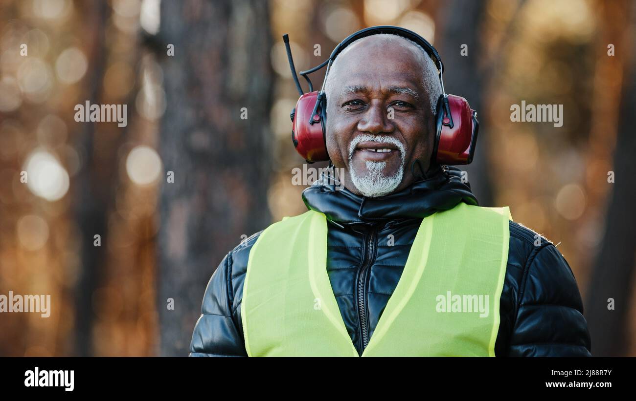 Älterer Mann professioneller Forstingenieur in schützenden schalldichten Kopfhörern, der im Wald steht und auf den kamerareifen afroamerikanischen Vorarbeiter blickt Stockfoto