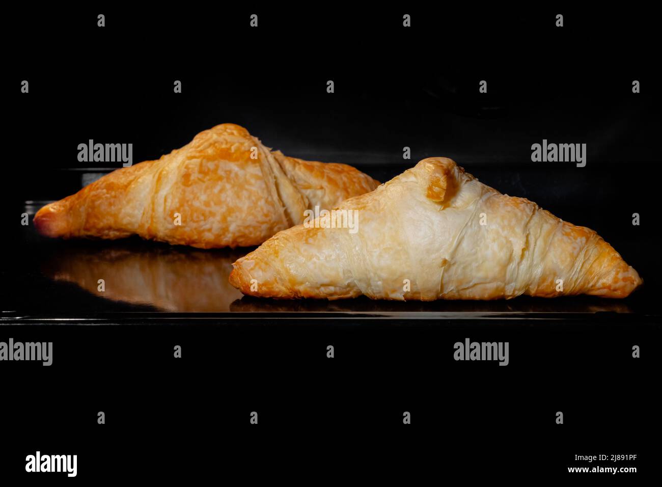 Zwei gebackene Croissants auf Tablett im elektrischen Ofen zu Hause, schwarzer Hintergrund Stockfoto