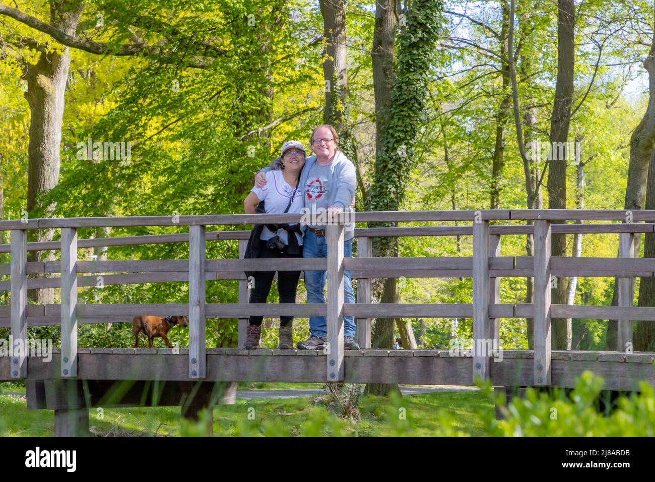 Wanderer reifes Paar lächelnd und glücklich auf einer Holzbrücke mit ihrem Dackel, Blick auf die Kamera, üppig grüne Bäume im Hintergrund, sonnigen Frühling Stockfoto