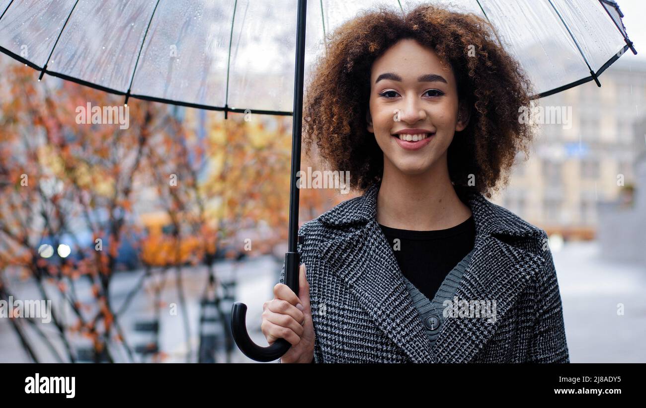 Lächelnd fröhlich afroamerikanisch 20s Modell Tourist girl Ausländer Frau mit transparentem Regenschirm in der Stadt stehend genießen regen reisen in neu Stockfoto