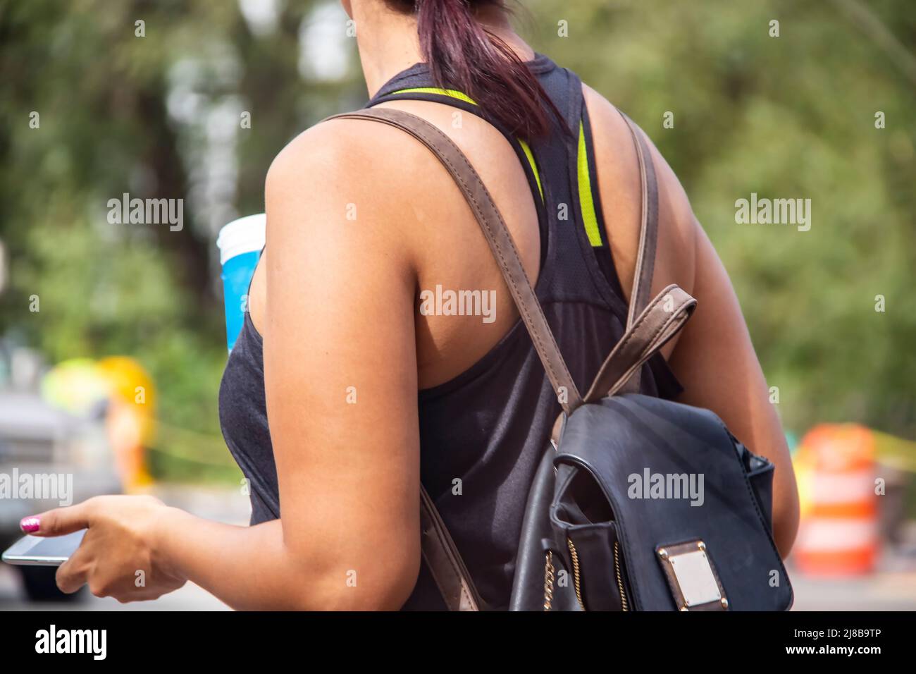Gekürzte, nicht wiedererkennbare Frau mit Sommer-T-Shirt und braunem und schwarzem Lederrucksack für Softdrinks und Handy vor Bokeh-Hintergrund Stockfoto