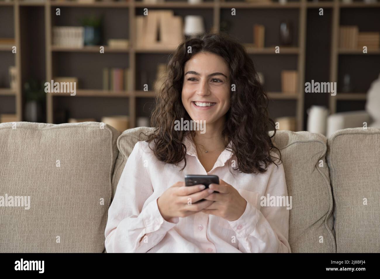 Frau sitzt auf dem Sofa und Smartphones starren zur Seite und sehen glücklich aus Stockfoto