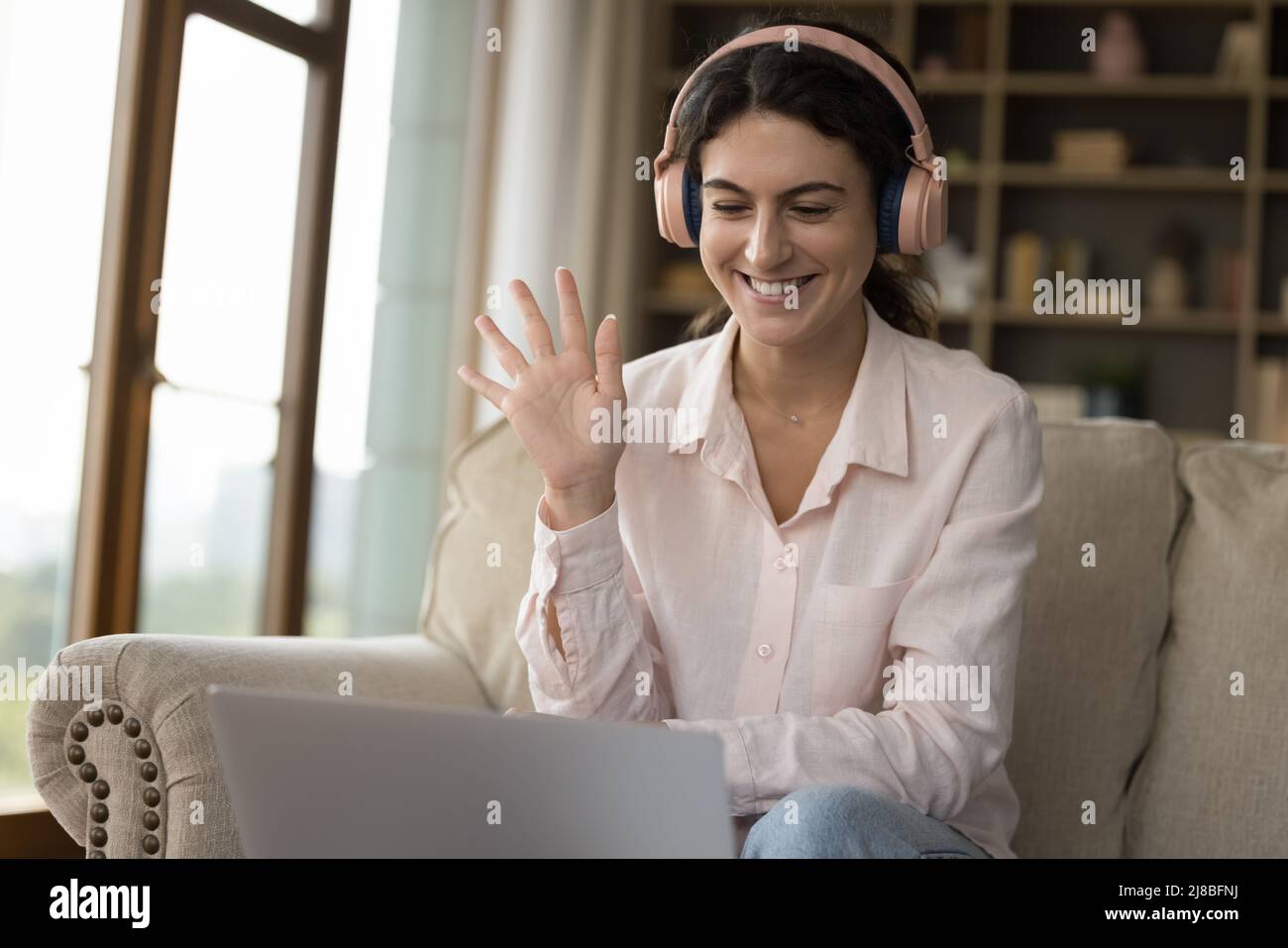 Eine Frau mit Kopfhörern starrt auf den Laptop und begrüßt einen Freund, der eine Videokonferenz startet Stockfoto