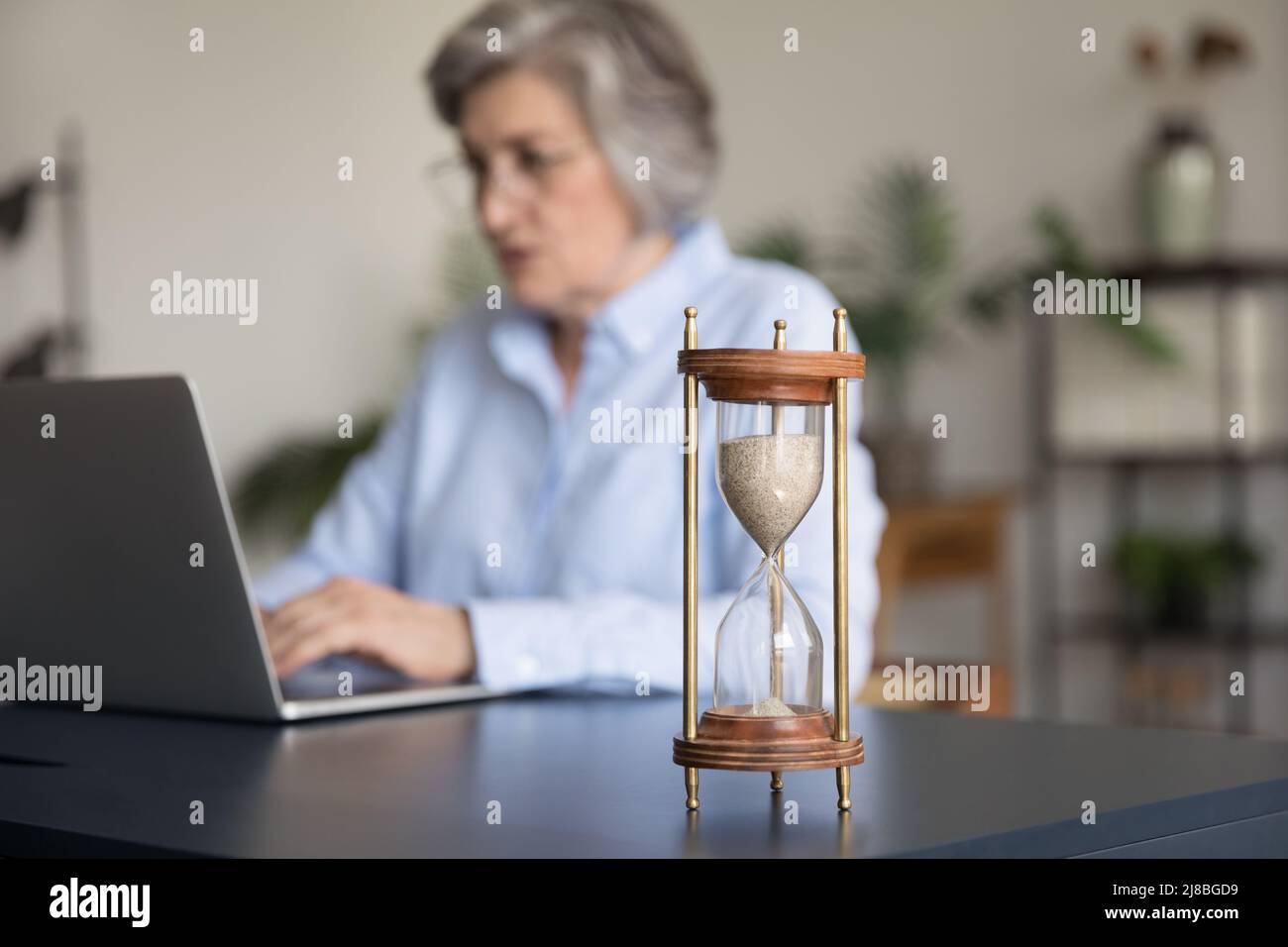 Fokussierte ältere, grauhaarige Geschäftsfrau, die an der Sanduhr einen Laptop verwendet Stockfoto