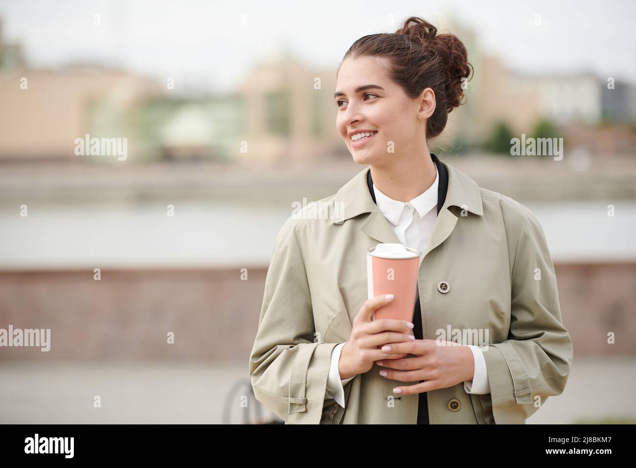 Lächelnde, unbeschwerte junge Geschäftsfrau in stilvollem Mantel, die während eines Spaziergangs durch die Stadt Kaffee trinkt Stockfoto