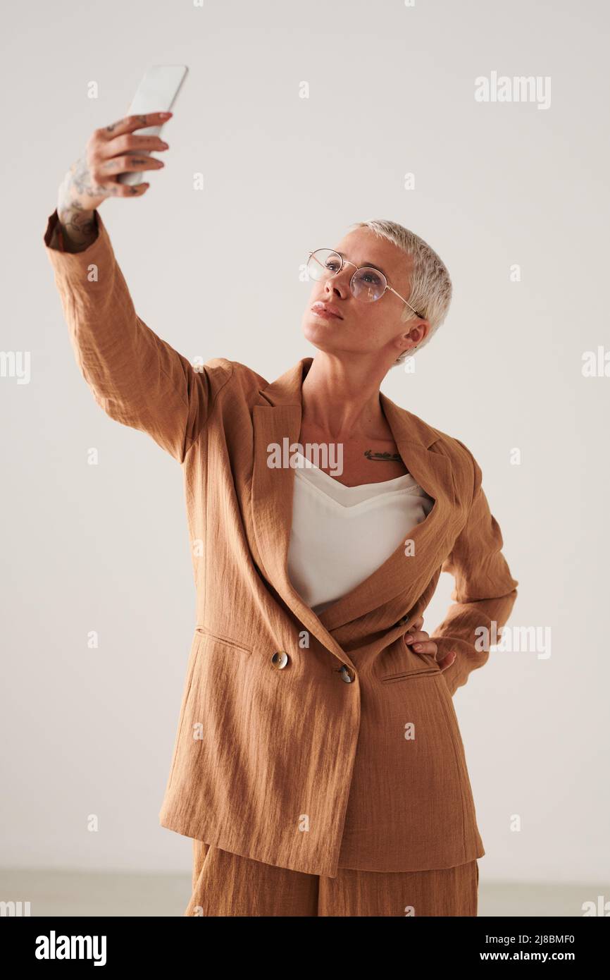 Stilvolle Geschäftsfrau mittleren Alters in lockeren Anzug nimmt Selfie auf dem Smartphone vor weißem Hintergrund Stockfoto