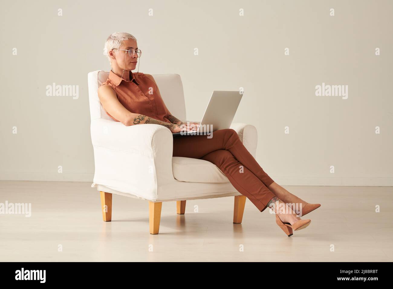 Freiberufliche Frau mittleren Alters in stilvollem Outfit sitzt in einem bequemen Sessel und mit Laptop, während sie an einem Online-Projekt arbeitet Stockfoto