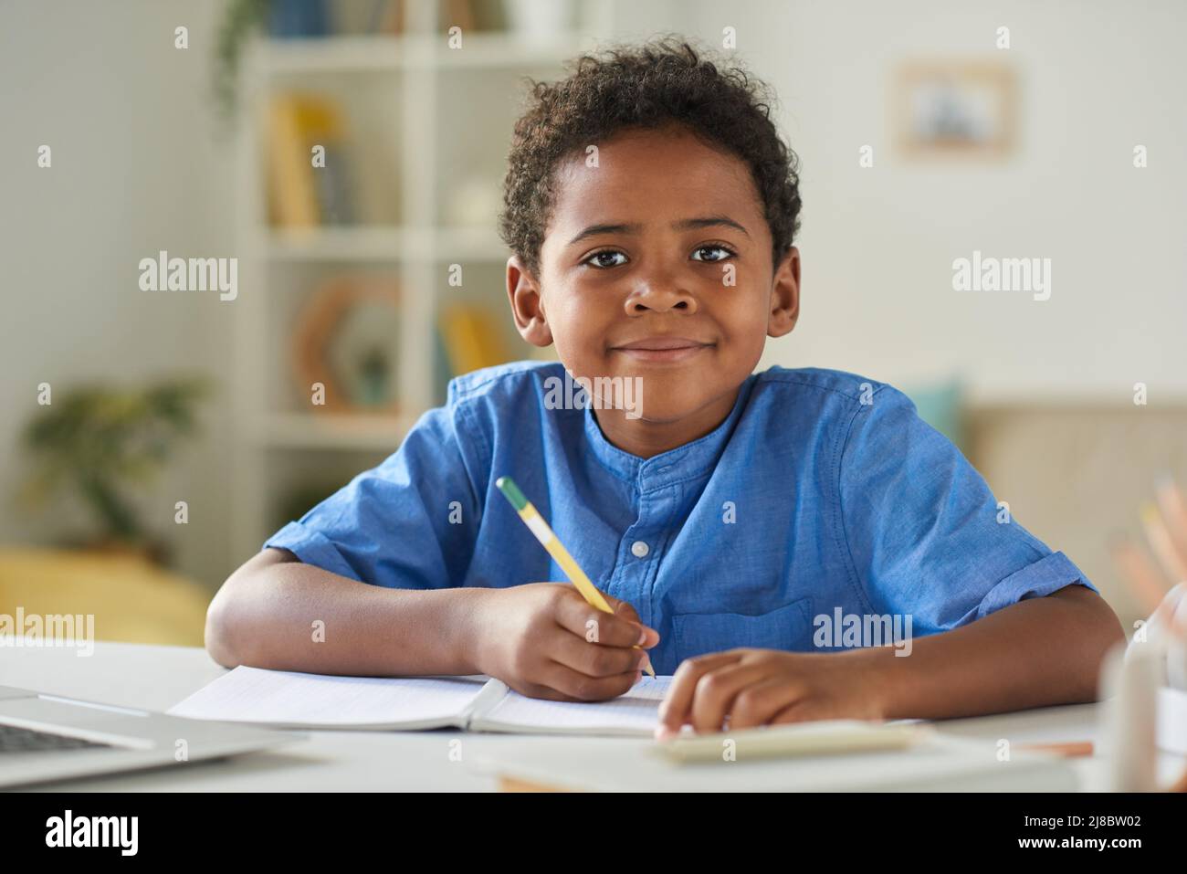 Porträt eines lächelnden afroamerikanischen Jungen in blauem Hemd, der am Tisch sitzt und Notizen im Arbeitsbuch macht Stockfoto