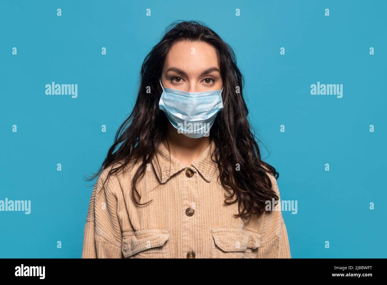 Serious junge europäische Brünette Frau in Schutzmaske Blick auf Kamera, isoliert auf blauem Hintergrund Stockfoto