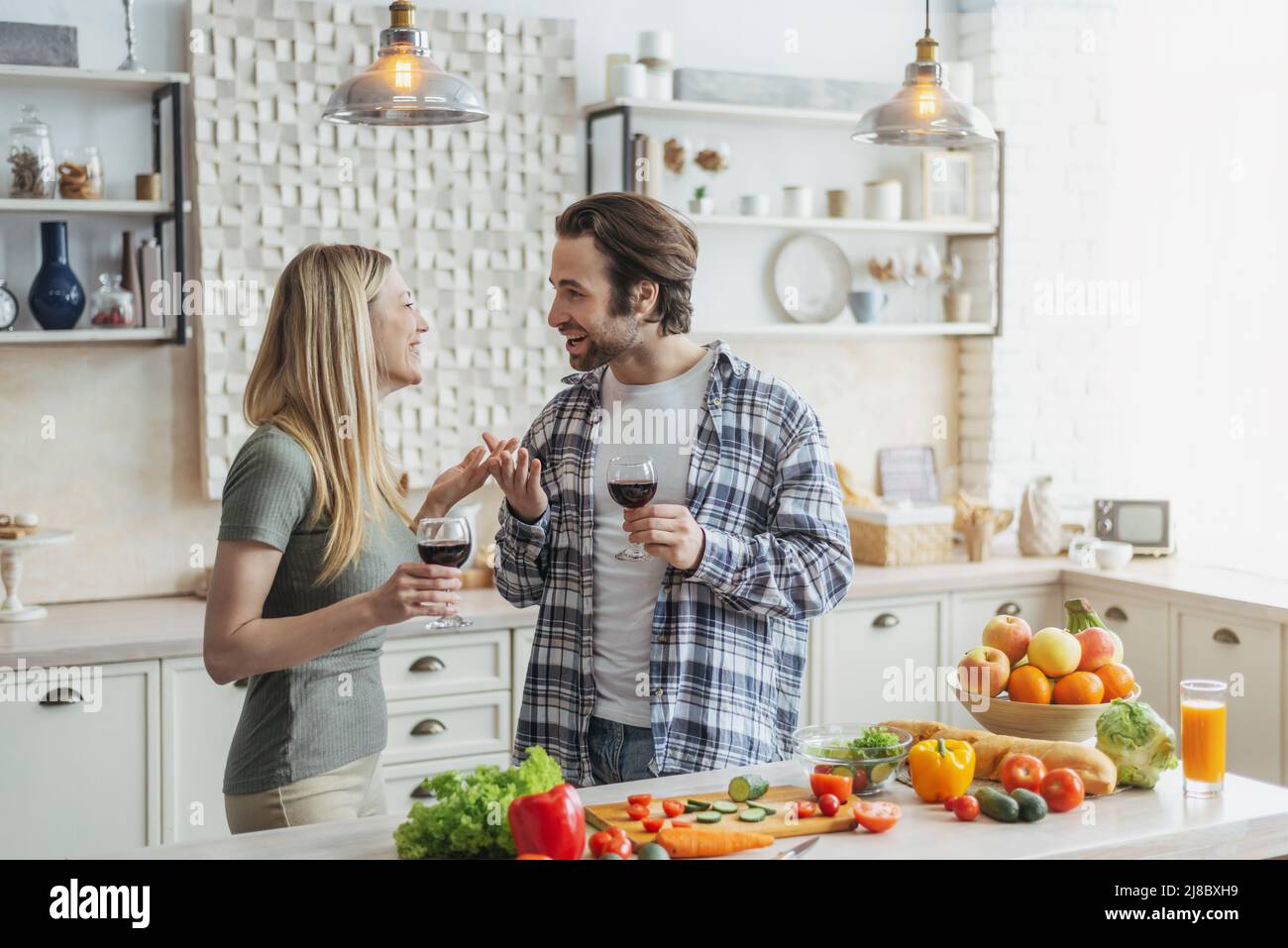 Zufriedene junge europäische Familie spricht, klirrende Gläser Wein am Tisch mit Gemüse in der modernen Küche Stockfoto