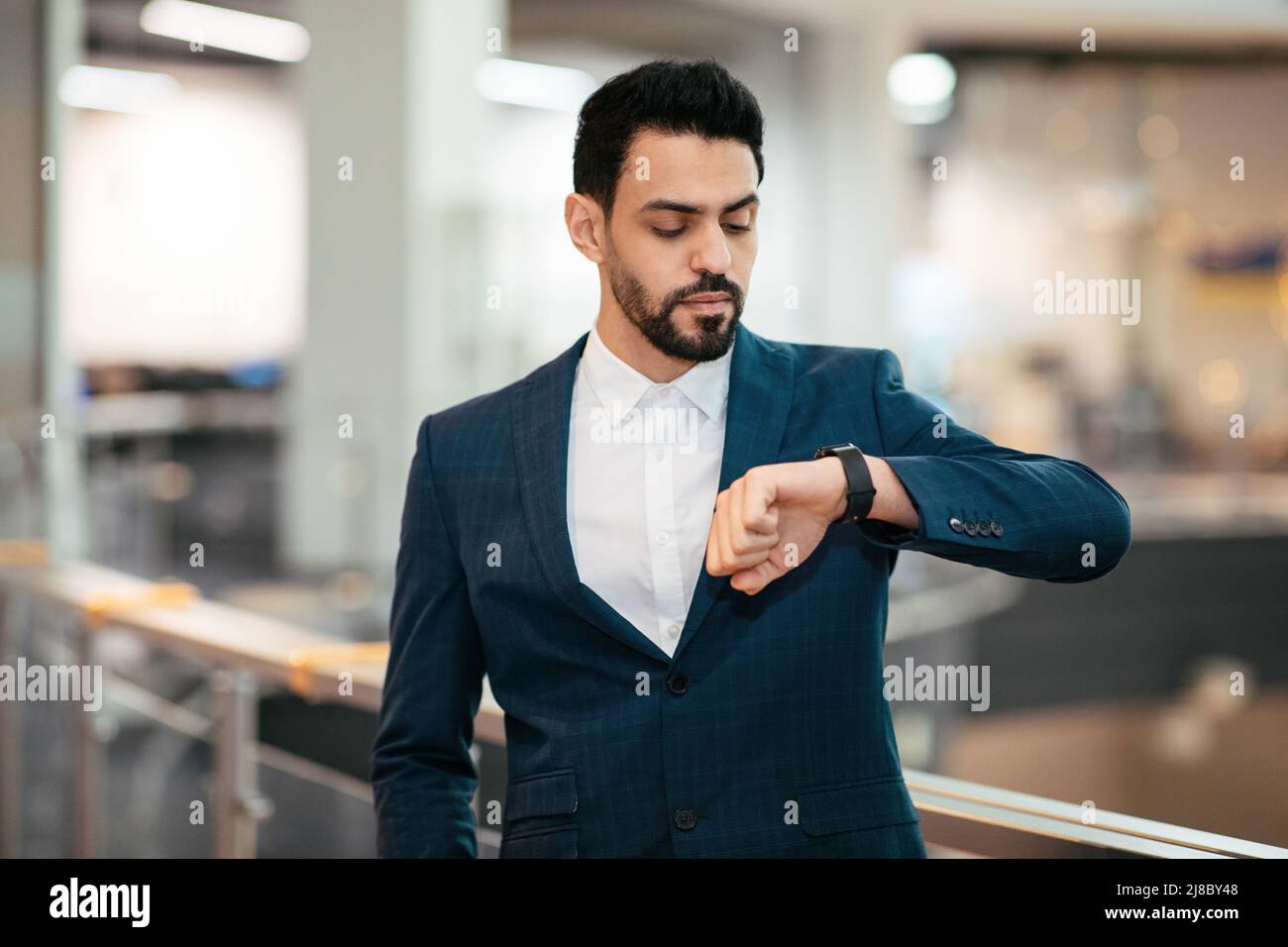 Serious ruhig attraktiven jungen arabischen ceo Mann mit Bart im Anzug sieht auf intelligente Uhr warten Client im Büro Interieur Stockfoto