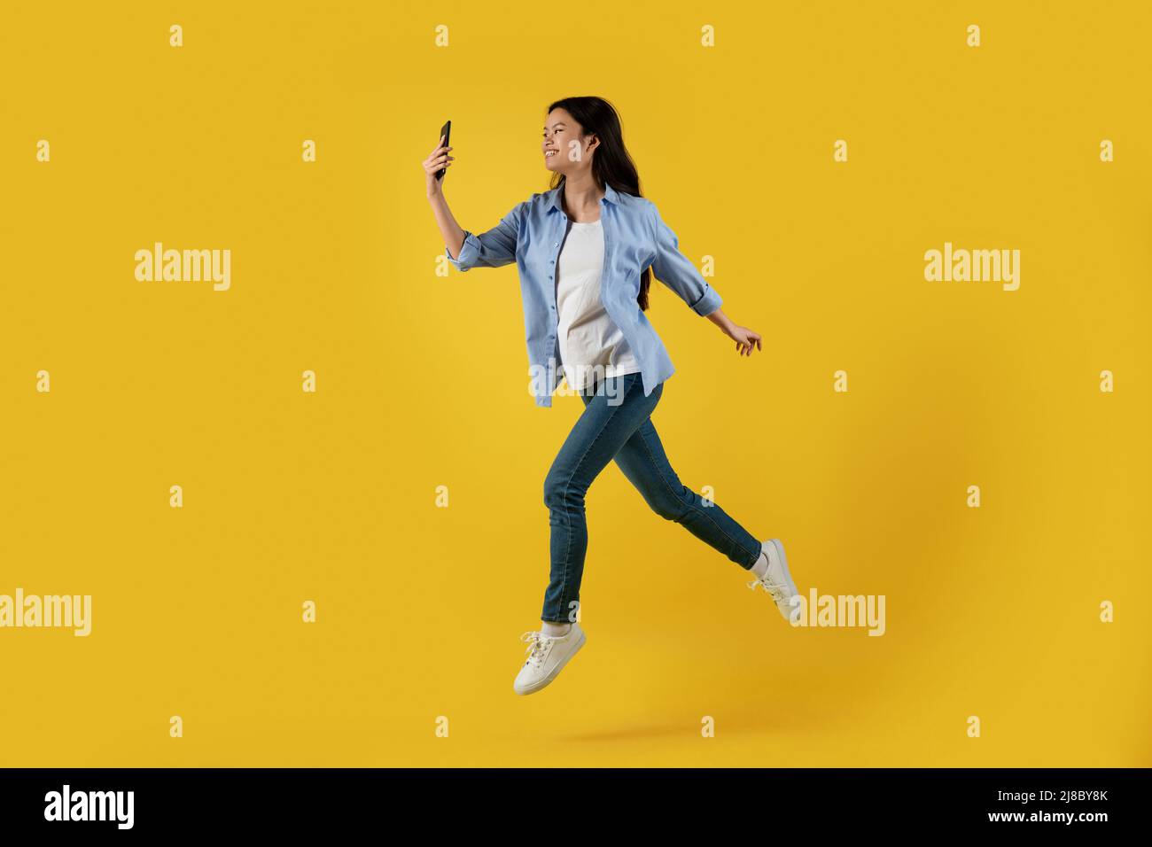 Frohes, tausendjähriges asiatisches Weibchen erstarrte im Luftspringen und betraf das Smartphone, isoliert auf gelbem Hintergrund Stockfoto