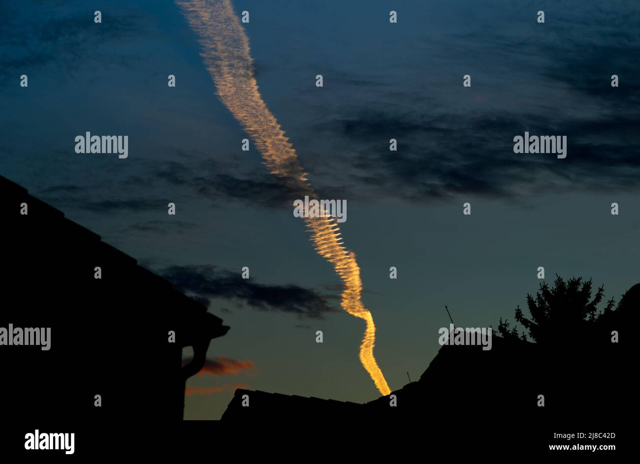 Einzelne ansprechende und leuchtende Kondensstreifen in helloranger Farbe über den Silhouetten der Dächer im dunkelblauen Abendhimmel (+ Haus) Stockfoto