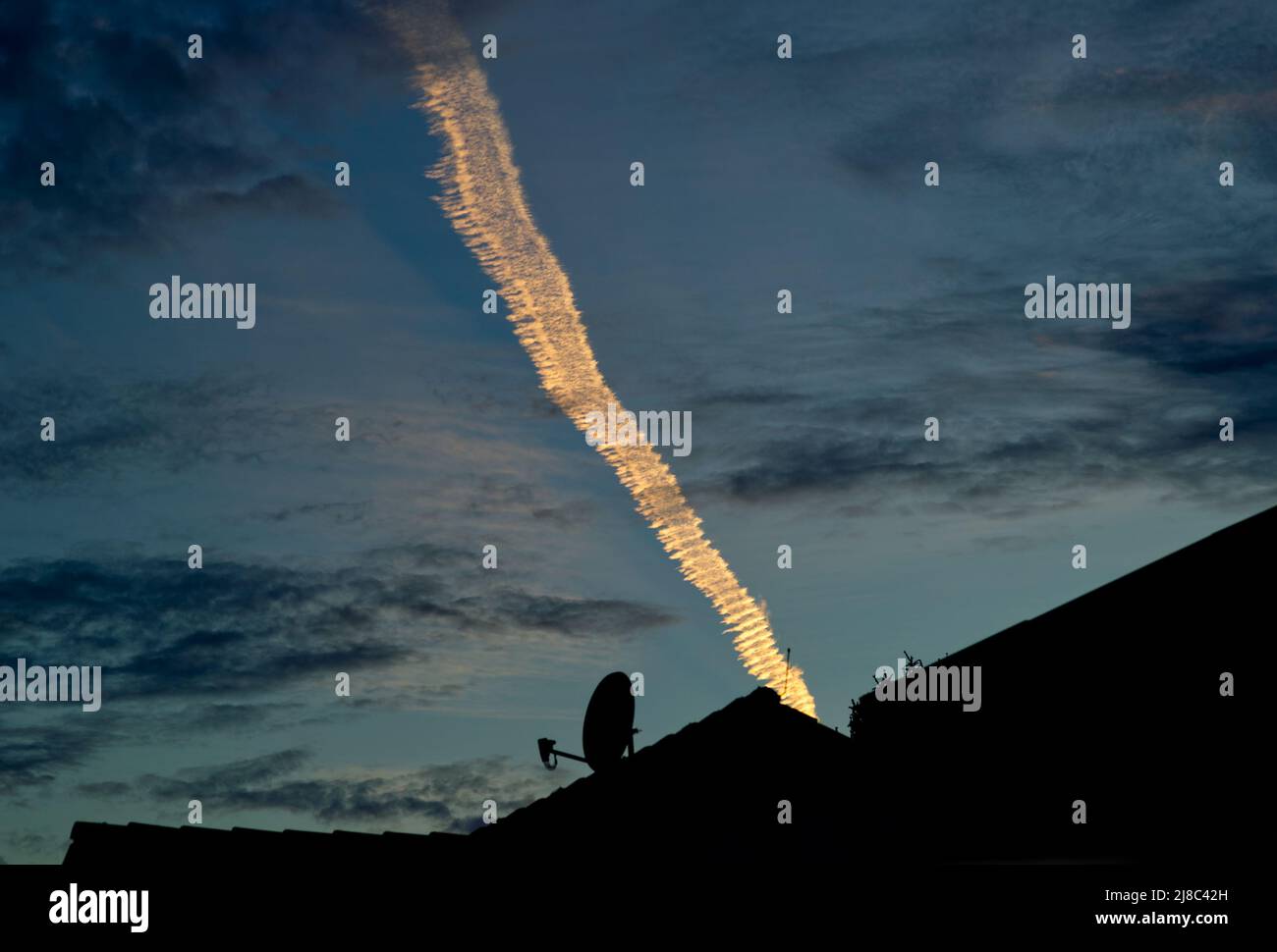Einzelne ansprechende und leuchtende Kondensstreifen in helloranger Farbe über den Silhouetten der Dächer im dunkelblauen Abendhimmel (Satellitenschüssel) Stockfoto