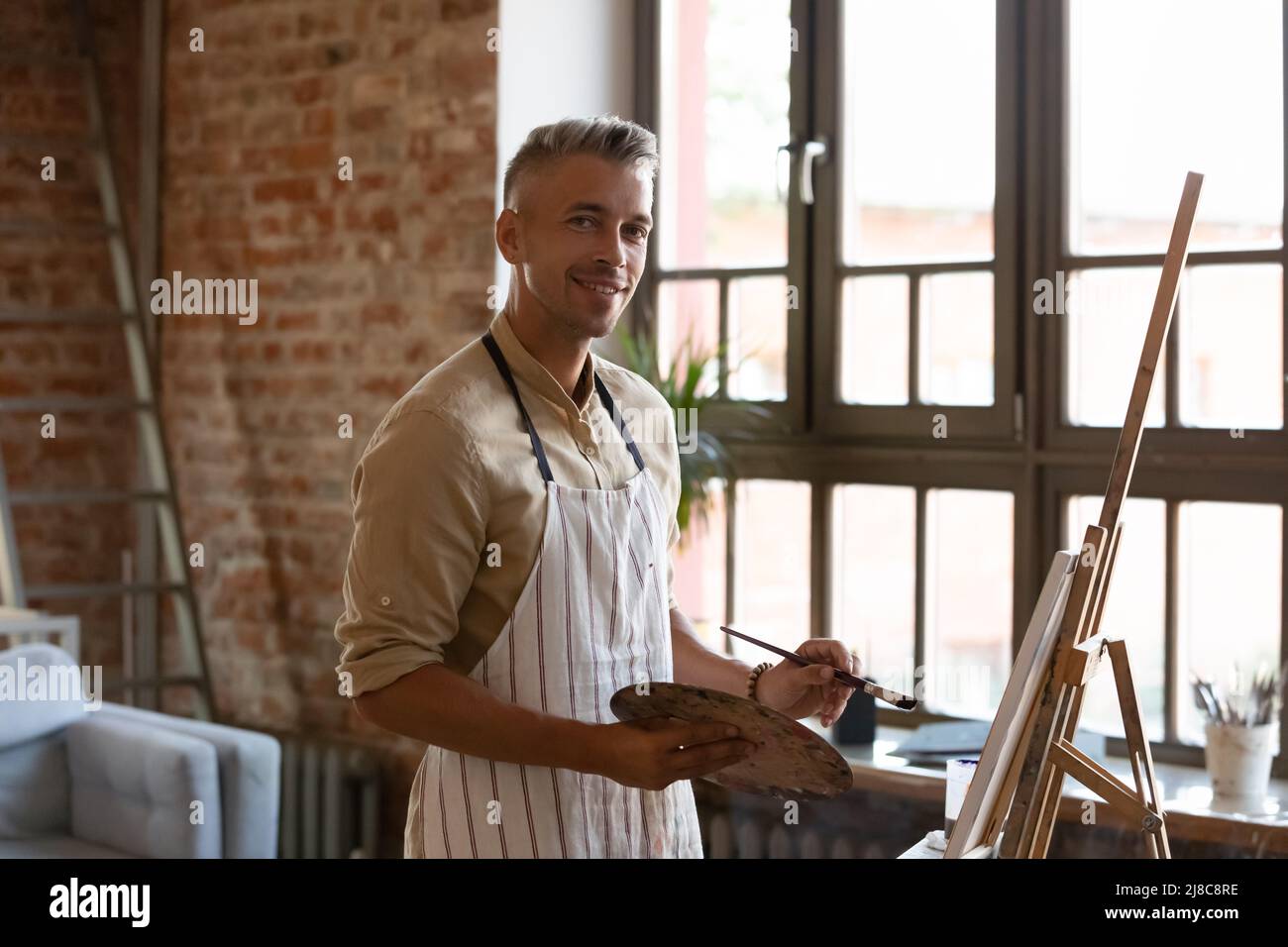 Hübscher Künstler, der Bilder auf Leinwand malt, posiert in der Werkstatt Stockfoto