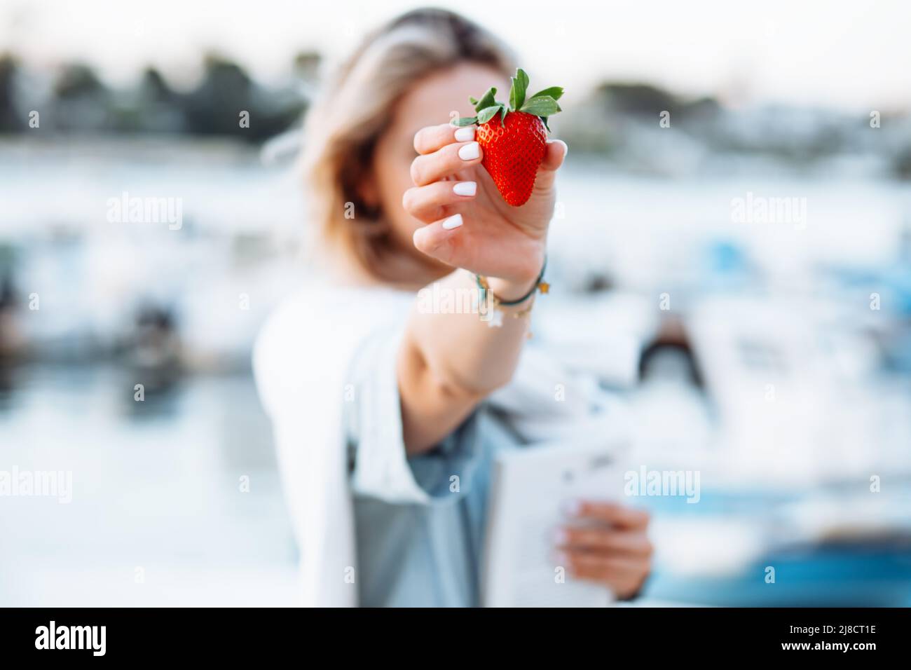 Porträt einer jungen Frau, die in der Hand reife, leckere Erdbeeren auf verschwommenem Hintergrund in Nahaufnahme hält und mit Booten und Yachten im Resort entlang des Uferdamms geht Stockfoto