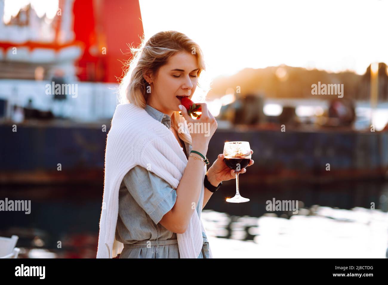 Porträt einer entspannenden Frau, die in einem roten Weinglas in der Hand hält und bei Sonnenuntergang frische Erdbeeren auf einem verschwommenen Hintergrund am Pier am Abend isst. Wandern am Meer entlang Stockfoto