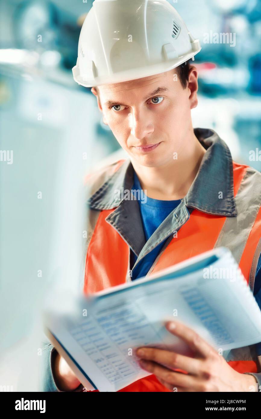 Der junge Inspektor oder Ingenieur in Helm und Bauweste führt Journaleinträge in der Produktionsanlage durch. Qualitätskontrolle der Arbeiten. Porträt eines kaukasischen Arbeiters im Zimmer. Stockfoto